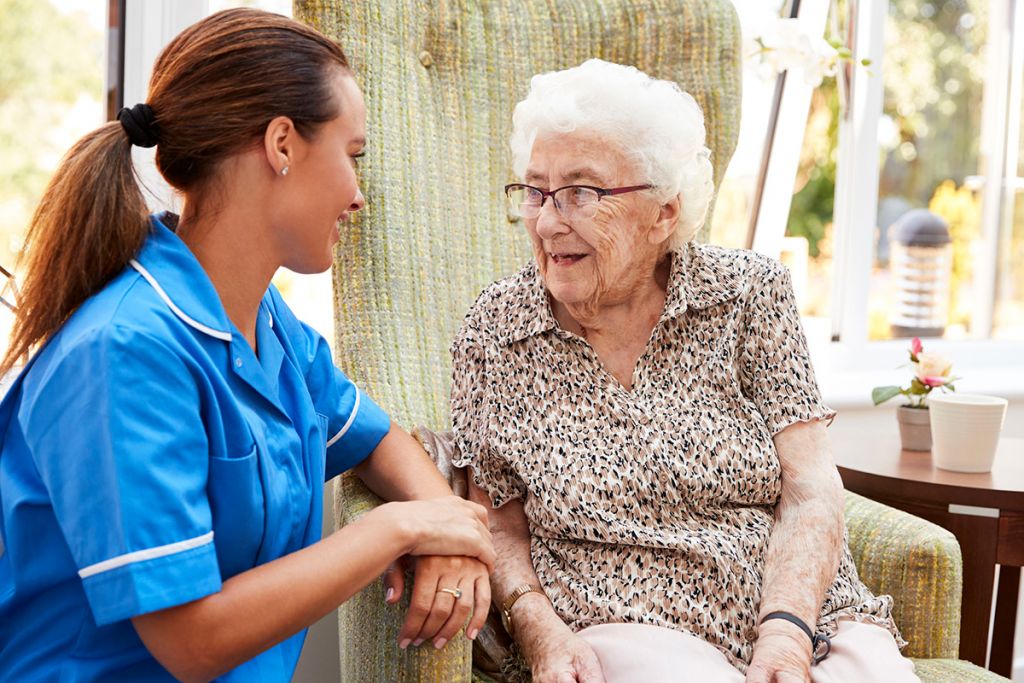 Senior-Woman-Sitting-In-Chair-And-Laughing-With-Nurse-In-Retirement-Home